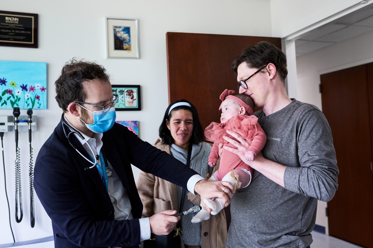 a dad holding a baby while a doctor checks her foot while a woman watches
